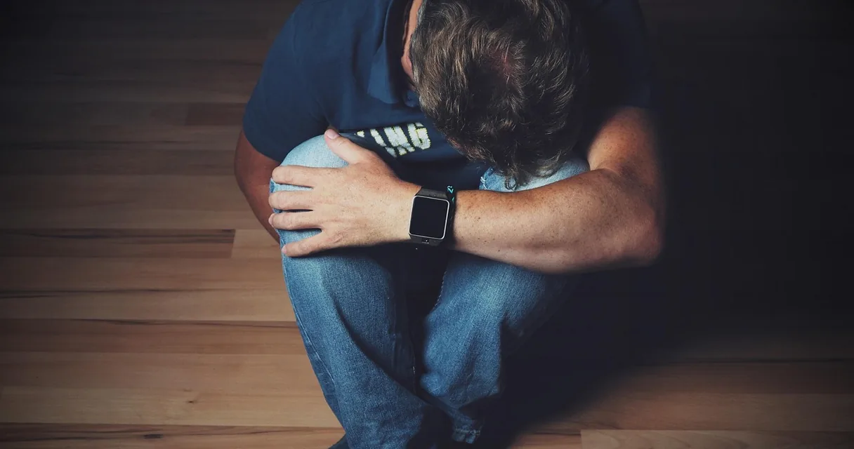 Homem sentado no chão abraçando os joelhos com a cabeça baixa, o que significa ser adicto