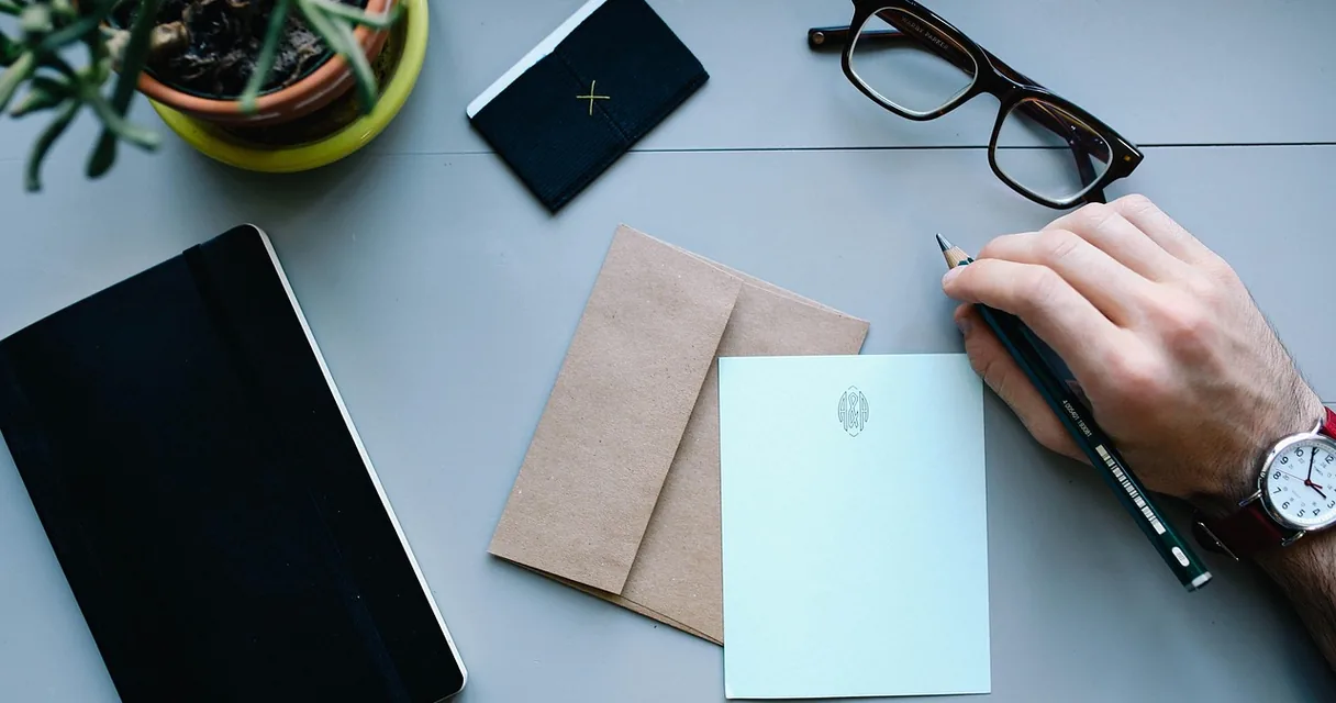 Mão escrevendo em um cartão ao lado de envelope e óculos sobre a mesa, carta para um dependente químico