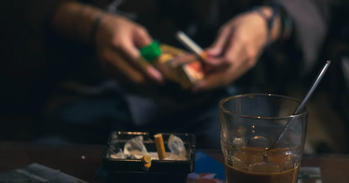 Mãos acendendo um cigarro ao lado de um copo de bebida e cinzeiro sobre a mesa, exemplos de drogas lícitas