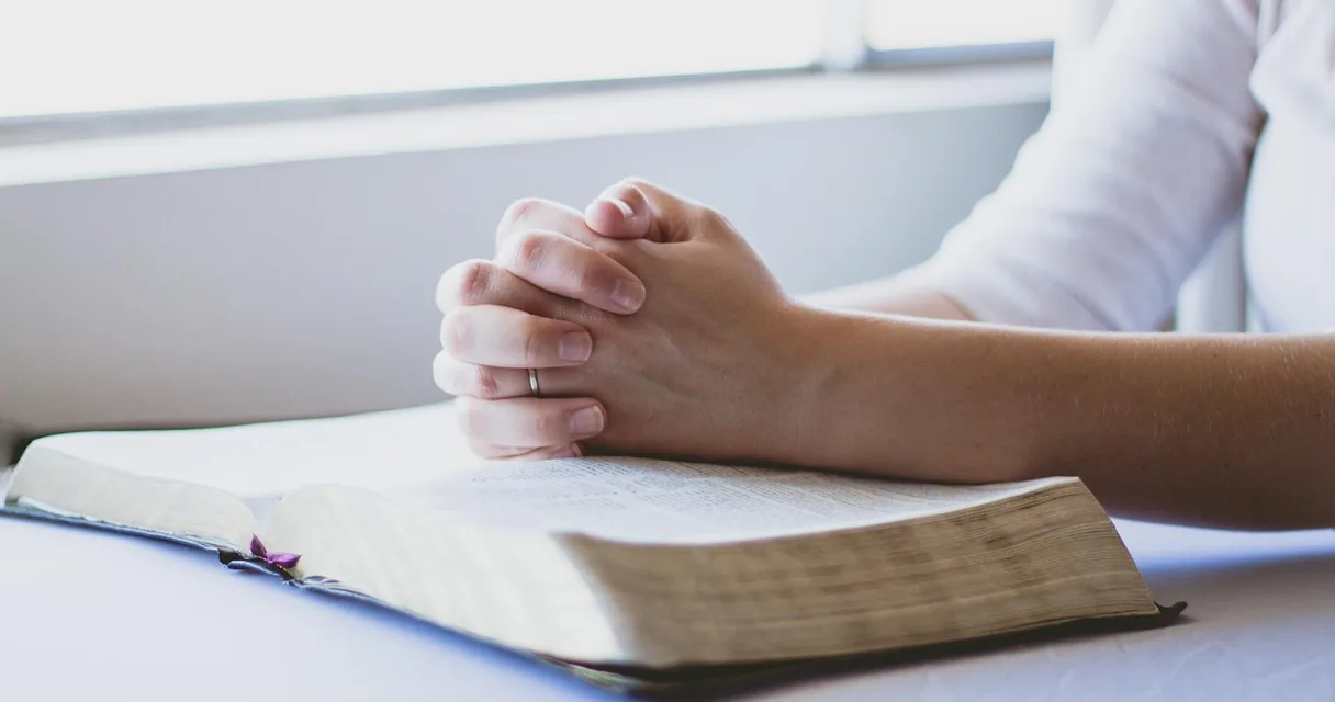 Mãos unidas em oração sobre uma bíblia aberta junto à janela, fé e espiritualidade na recuperação